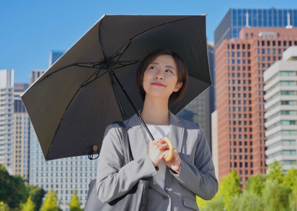 日差しの強い都会の街並みで、黒い日傘を差して微笑むスーツ姿の女性。