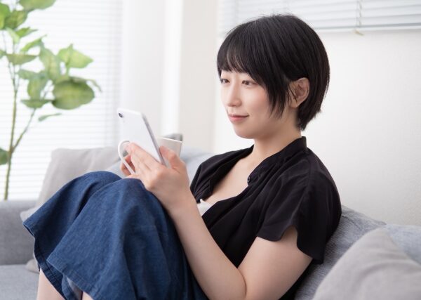 若い女性がリラックスしてソファに座り、スマートフォンを手に微笑んでいる。