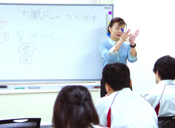教室で生徒たちに向かって熱心に説明しているタニカワ久美子講師。ホワイトボードには「健康」に関する日本語のキーワードと図が描かれている。