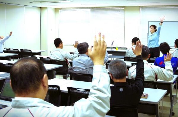 講師が教室で多くの人々に向かって話している。参加者は手を挙げて講師に注目している。